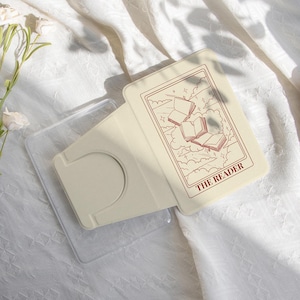 May include: A cream-colored phone card holder with a clear plastic case. The card holder features a design of open books and the text "THE READER" in a reddish-brown color. The background is a white, textured fabric with a floral arrangement.
