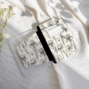 Può includere: Un piccolo quaderno con un motivo floreale di fiori di iris bianchi e neri. Il quaderno è aperto, rivelando pagine nere, e si trova in una custodia di plastica trasparente. La copertina recita "COMPOSITION BOOK".
