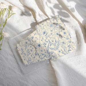 以下が含まれることがあります： クリーム色と青色の花柄のマスクとケース。クリーム色の背景に青い花と葉のデザインが施されています。「happylifeenjoydesign」の文字が見えます。白いテクスチャード加工された布の上に置かれています。