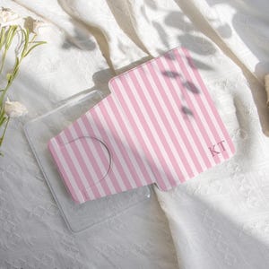 Puede incluir: Una funda de teléfono a rayas rosas y blancas, con las iniciales "KT", se muestra abierta, revelando un soporte transparente para el teléfono. La funda está sobre una superficie blanca texturizada. Las palabras "happylifeenjoydesign" están en la parte inferior.