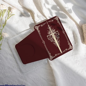 Puede incluir: Un tarjetero burdeos con un diseño de espada dorada y borde decorativo. El tarjetero está colocado en una funda de plástico transparente. El texto "happylifeenjoydesign" está en la parte inferior izquierda.