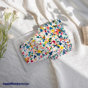 Può includere: Una custodia trasparente per telefono con un design floreale colorato. La custodia presenta un motivo di piccoli fiori blu, rosa, gialli e verdi su sfondo bianco. Il testo "happylifeenjoydesign" è visibile in basso.