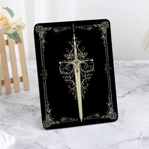 May include: A black rectangular item featuring a detailed, cream-colored illustration of a sword with ornate embellishments. The design is centered and framed by decorative flourishes.