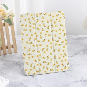 Può includere: Una custodia per tablet color crema con un motivo ripetuto di piccoli fiori gialli e foglie verdi. La custodia è in piedi su una superficie di marmo, mostrando il suo design floreale.