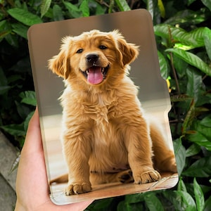 Puede incluir: Una funda para tableta con la imagen de un cachorro de golden retriever sonriente con la lengua fuera. La funda tiene un fondo marr&oacute;n y el texto "happylifeenjoydesign" en blanco. La funda se sostiene contra un fondo de hojas verdes.
