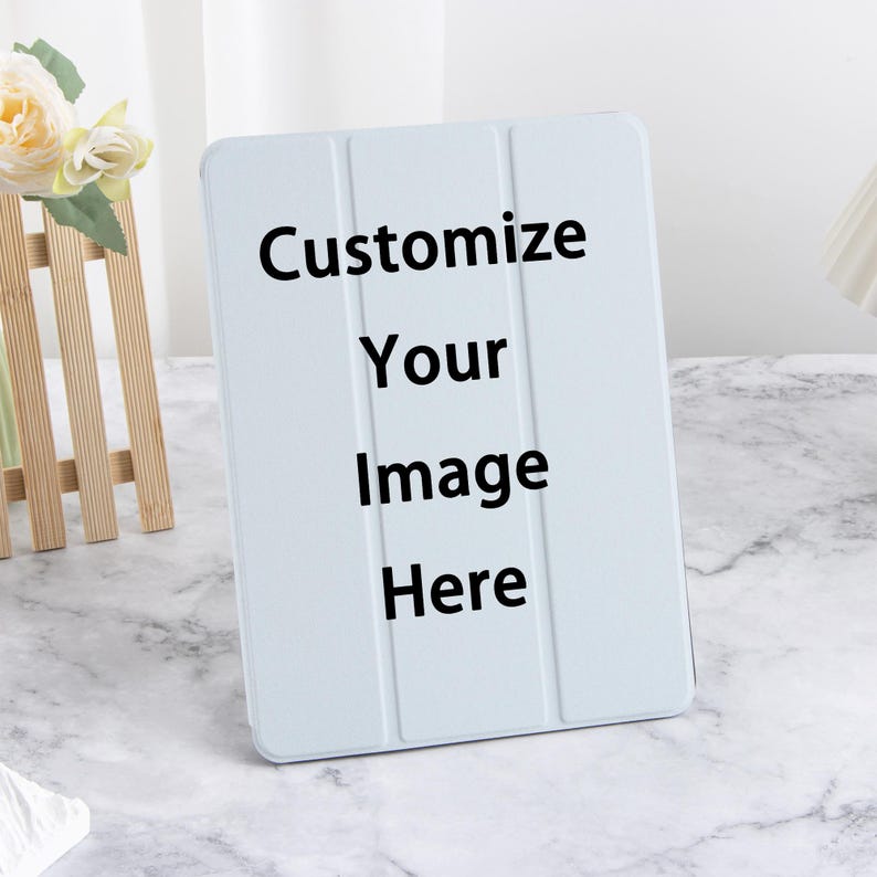 Może przedstawiać: Jasnoniebieskie etui na tablet z czarnym napisem "Customize Your Image Here". Etui ma potr&oacute;jną konstrukcję i stoi pionowo. Drewniany element dekoracyjny i kwiaty znajdują się w tle.