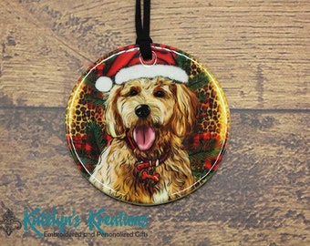 May include: A round ceramic Christmas ornament featuring a brown and white dog wearing a red Santa hat. The dog is surrounded by a red and black plaid pattern, a leopard print, and green pine branches.