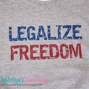Op de afbeelding: Een grijs T-shirt met de tekst "Legalize Freedom" gedrukt in blauwe en rode, verweerde letters.