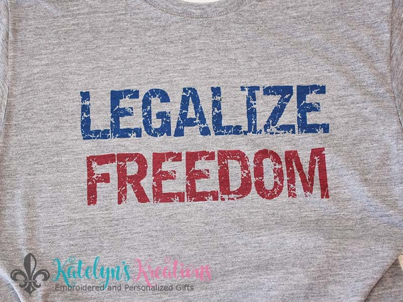 Op de afbeelding: Een grijs T-shirt met de tekst "Legalize Freedom" gedrukt in blauwe en rode, verweerde letters.