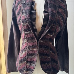 Peut inclure: Un blazer marron foncé avec un motif rayé texturé dans les tons bordeaux, noir et beige. Le blazer a des manches longues et deux boutons. La doublure est en satin marron foncé. Un collier ras du cou noir avec un pendentif est visible.