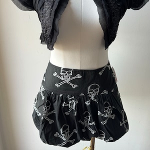 May include: A black cropped bolero with a ruffled neckline and short sleeves. The bolero is paired with a black mini skirt featuring a white skull and crossbones print.