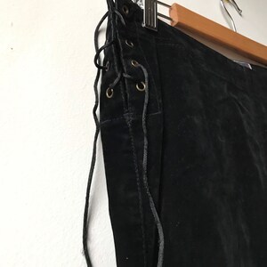 K&ouml;nnte beinhalten: Schwarzer Wildlederschurz mit Schn&uuml;rung an der Seite. Der Rock h&auml;ngt an einem Holzb&uuml;gel.