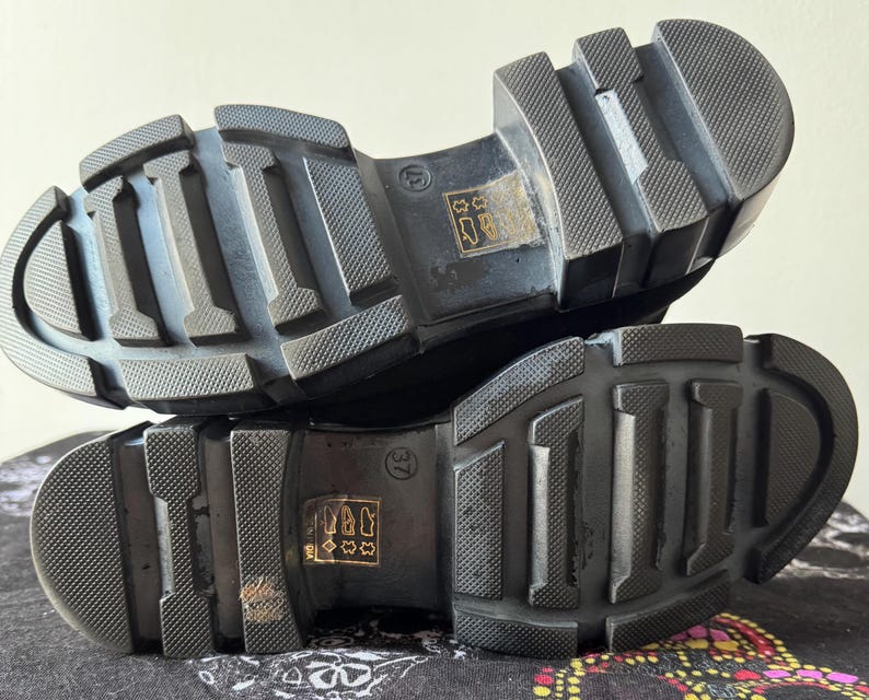 May include: Close-up of the black rubber soles of a pair of shoes. The soles have a textured tread pattern and are designed for grip and traction.