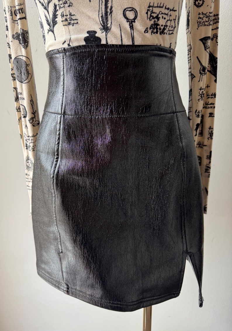 May include: A black faux leather mini skirt with a side slit. The skirt has a high waist and a fitted silhouette.