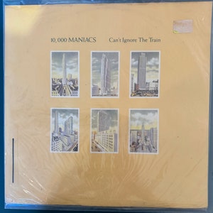May include: A vintage vinyl record album cover featuring a collage of six images of cityscapes with tall buildings. The album is titled "10,000 Maniacs" and "Can't Ignore The Train".