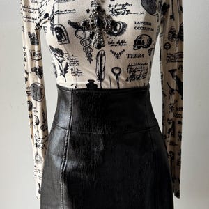May include: A white long-sleeved shirt with a black and white print of various symbols, including keys, a skull, and a hand with an eye. The shirt is paired with a black leather skirt.