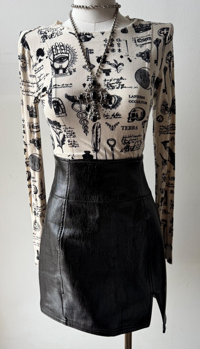May include: A white long-sleeved shirt with a black and white print of various symbols, including keys, a skull, and a hand with an eye. The shirt is paired with a black leather skirt.