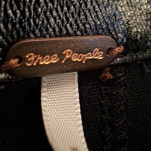 May include: A close-up of a brown leather tag with the words "Free People" embossed in gold lettering. The tag is attached to a black garment with a white ribbon.