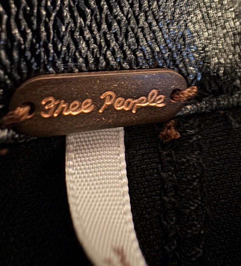 May include: A close-up of a brown leather tag with the words "Free People" embossed in gold lettering. The tag is attached to a black garment with a white ribbon.