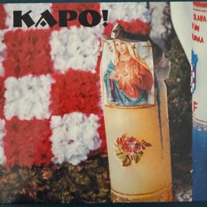 May include: A white candle with a red and white floral design and a picture of the Virgin Mary on the front. The candle is sitting on a red and white checkered surface. The text "KAPO!" is visible in the top left corner of the image.