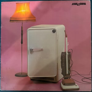 May include: Album cover featuring a cream-colored refrigerator, a floor lamp with an orange lampshade, and a vintage Hoover vacuum cleaner against a pink background. The text "THE CURE" is in the upper right corner.