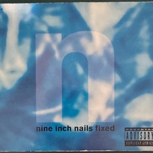 Puede incluir: Una portada de álbum de música cuadrada con un fondo azul y blanco borroso y una letra n minúscula grande en el centro. El texto nine inch nails fixed está impreso en negro en la parte inferior, junto a una etiqueta Parental Advisory en blanco y negro.