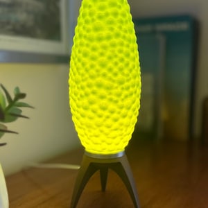 May include: A modern, glowing table lamp with a textured, yellow-green shade. The lamp sits on a dark gray tripod base. The lamp's shade has a unique, dimpled surface, and emits a soft, warm light, creating a decorative accent.