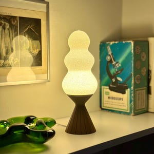 Puede incluir: Una lámpara de mesa decorativa con una forma apilada y orgánica. La lámpara tiene una pantalla texturizada de color blanco roto y una base marrón oscuro. La lámpara está encendida, emitiendo un brillo cálido. Una caja de microscopio vintage y una impresión enmarcada están en el fondo.