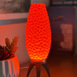 Puede incluir: Lámpara de mesa naranja brillante con una superficie texturizada con un patrón de puntos. La lámpara se apoya sobre una base de trípode con patas de color gris oscuro. Junto a la lámpara hay una maceta geométrica blanca con una planta suculenta.