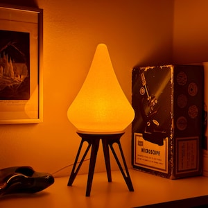 Puede incluir: Lámpara de mesa de estilo retro con una pantalla amarilla texturizada en forma de lágrima. La lámpara se apoya sobre una base trípode marrón oscuro. En el fondo se ven una impresión enmarcada y una caja con la etiqueta "Microscope".