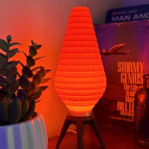 Puede incluir: Una lámpara de lava naranja brillante con un diseño texturizado y acanalado, montada sobre una base de trípode de madera oscura. La lámpara está encendida, emitiendo un brillo cálido. Una planta suculenta en maceta y álbumes de discos vintage están en el fondo.