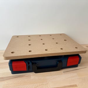 Peut inclure: Un établi en bois brun clair avec plusieurs trous espacés, reposant sur une boîte à outils bleu foncé avec des loquets rouges. La boîte à outils a une poignée noire et la marque "Bosch" est visible. Deux goupilles métalliques sont également présentes.