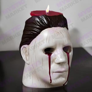 K&ouml;nnte beinhalten: Eine wei&szlig;e Kerze in Maskenform mit dunklem Haar und roten Wachstr&auml;nen. Obenauf befindet sich eine brennende rote Kerze. Ein dekorativer Artikel zum Thema Halloween.