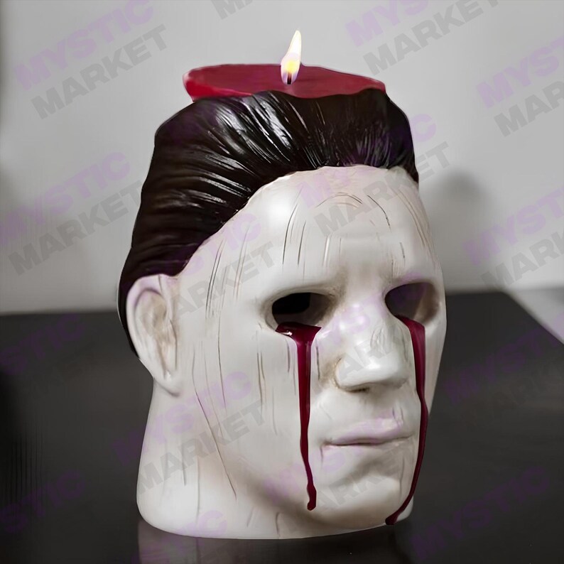 K&ouml;nnte beinhalten: Eine wei&szlig;e Kerze in Maskenform mit dunklem Haar und roten Wachstr&auml;nen. Obenauf befindet sich eine brennende rote Kerze. Ein dekorativer Artikel zum Thema Halloween.