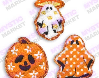May include: Three felt Halloween patches: a pumpkin with a smiling face and white flowers, a ghost with orange and white polka dots, and a mushroom with a white ghost inside and gold glitter.