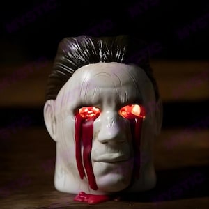 May include: A white Halloween head decoration with black hair. The eyes glow red, and red liquid appears to be dripping from them. The head is on a wooden surface.