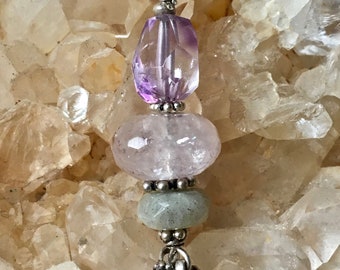 May include: A silver chain necklace with a pendant featuring three gemstone beads. The top bead is a faceted purple amethyst, the middle bead is a clear quartz, and the bottom bead is a green aventurine. The pendant is accented with silver beads and charms.