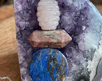 May include: A silver necklace featuring a pendant composed of a light pink, carved stone, a pink and gray crystal, and a blue lapis lazuli stone. The pendant is displayed against a purple amethyst geode.
