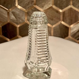May include: A clear glass salt shaker with a textured, faceted design. The shaker has a round top and a hexagonal base.