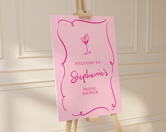 May include: Pink bridal shower welcome sign with a pink cursive font that says "Welcome to Stephanie's Bridal Shower". A pink martini glass with olives is above the text.