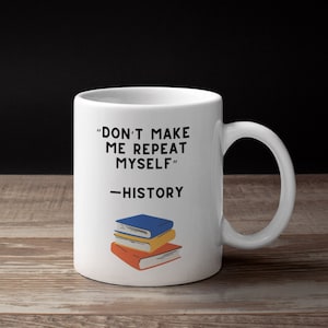 May include: White ceramic mug with the text "DON'T MAKE ME REPEAT MYSELF - HISTORY" in black. Below the text are illustrations of three stacked books in blue, yellow, and orange. The mug is on a wooden surface.