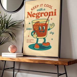 以下が含まれることがあります： 「KEEP IT COOL with a Negroni」の文字が入った、フレーム入りのヴィンテージスタイルのポスター。イラストは、笑顔の顔、オレンジスライス、ストローが付いた漫画のネグローニカクテルです。ポスターには、材料として25mlのカンパリ、ジン、ベルモットが含まれています。