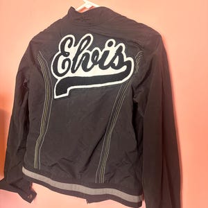 May include: A black jacket featuring a large white and black embroidered patch on the back with the text Elvis in a script font. The jacket includes vertical yellow stitching details, a stand-up collar, and a grey horizontal stripe at the hem.