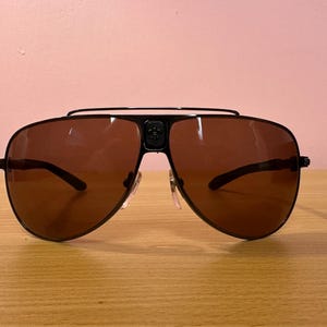 May include: Black aviator sunglasses with brown tinted lenses. The sunglasses have a black metal frame with a double bridge and a decorative emblem on the center. The lenses are a dark brown color.