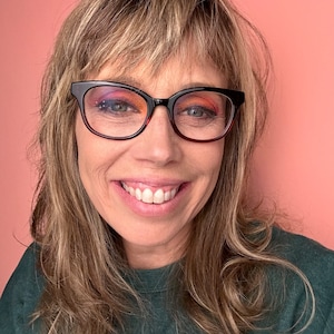 Könnte beinhalten: Eine Frau mit langen braunen Haaren, die einen dunkelgrünen Pullover und eine schwarze Brille mit orangefarbenen Gläsern trägt. Sie lächelt.