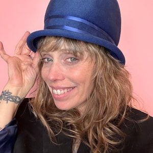 Puede incluir: Una mujer con un sombrero de fieltro azul de ala ancha y una cinta azul. Está sonriendo y tiene el pelo largo y ondulado rubio.