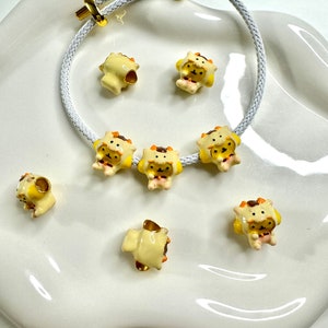 May include: A white cord bracelet with gold clasps and yellow ceramic animal charms. The charms are shaped like a bear wearing a yellow and orange hat.