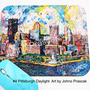 May include: A colorful mousepad featuring an abstract cityscape painting with buildings in shades of orange, purple, and blue. The mousepad has a blue border and the text "#4 Pittsburgh Daylight Art by Johno Prascak". A light blue wireless mouse is in the foreground.