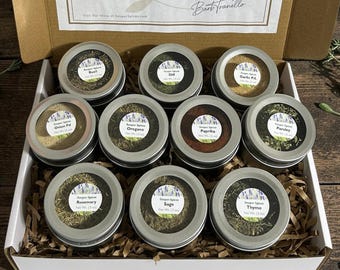May include: A cardboard gift box containing nine round metal tins filled with various dried herbs and spices, including basil, dill, garlic powder, onion powder, oregano, paprika, parsley, rosemary, sage, and thyme, each with a clear lid.