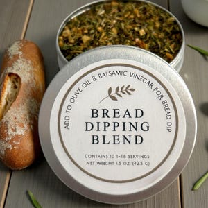 May include: A silver tin of bread dipping blend with the text "Bread Dipping Blend". The tin is open, revealing a mix of herbs and spices. A loaf of bread is to the left. The label says "Add to Olive Oil & Balsamic Vinegar for Bread Dip".